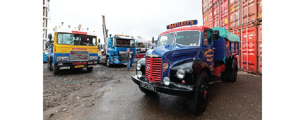 A collection of vintage and classic trucks is parked in an industrial yard with shipping containers in the background. A restored blue and black truck with "Bartlettās" branding is prominently featured in the foreground, while other colorful classic trucks, including a yellow and blue one, are visible behind it. A man is seen walking among the trucks.