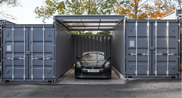 The image shows a black car parked under a canopy connecting two shipping containers on either side. The canopy has a translucent covering, allowing natural light to enter. The containers are closed, creating a symmetrical arrangement with the car centered beneath the canopy. In the background, trees with yellow and green foliage are visible, suggesting an outdoor setting. The scene showcases a secure and organised container-based parking or storage solution.