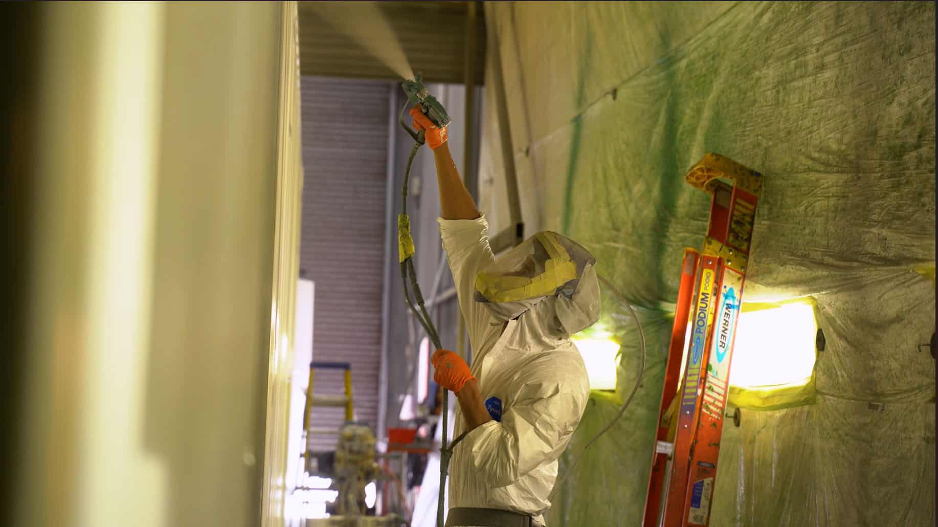 A worker in protective gear, including a full-body suit, gloves, and a hood with a face covering, is spray-painting a large shipping container inside a paint booth. The worker is holding a spray gun high above their head, applying an even coat of paint. The surrounding area is covered in protective plastic sheeting, with bright lights illuminating the workspace. A red ladder and various equipment are visible in the background.