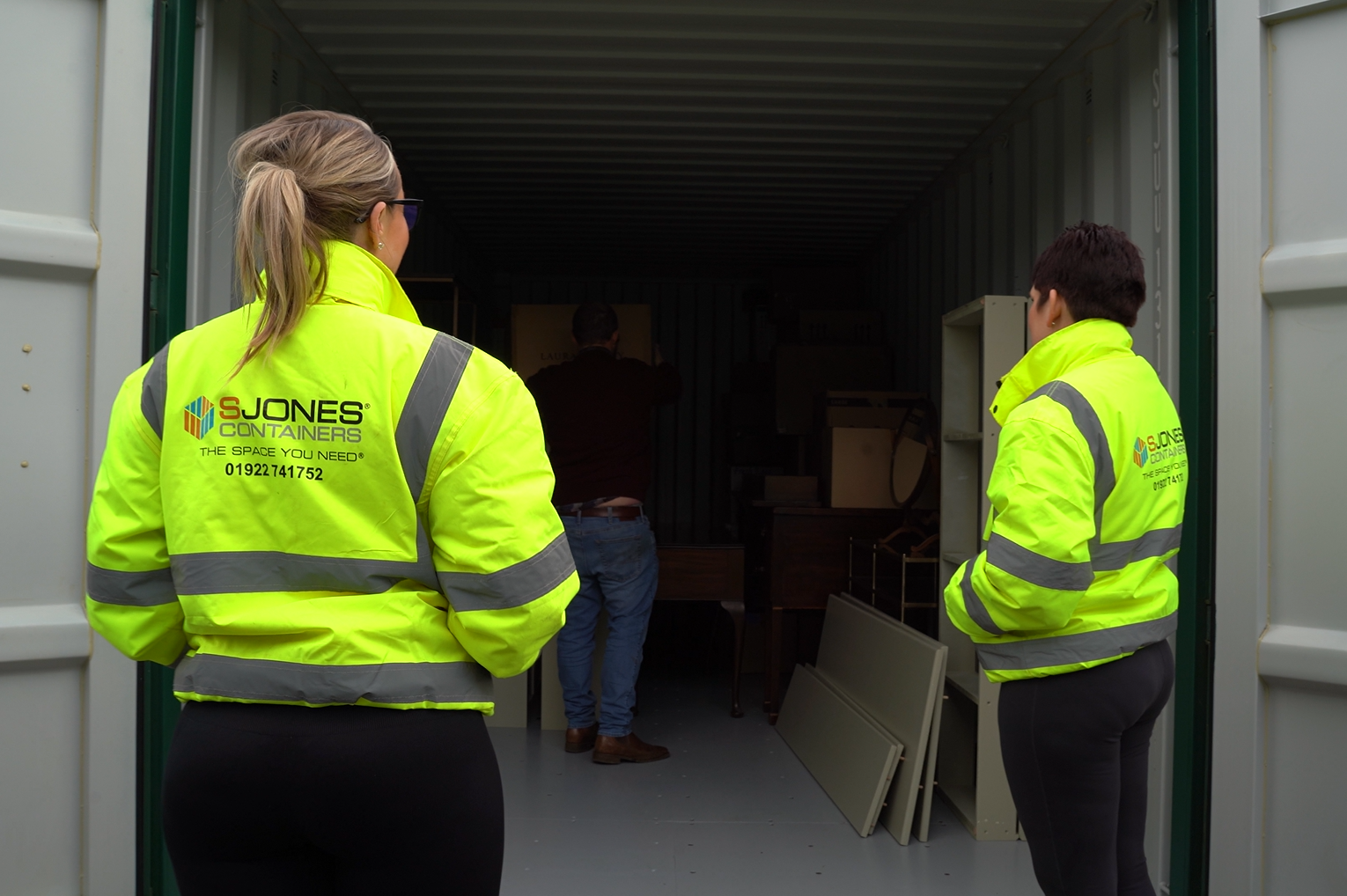 Two individuals in high-visibility yellow jackets with reflective stripes are standing at the entrance of an open storage container, looking inside. The jackets display the logo for 