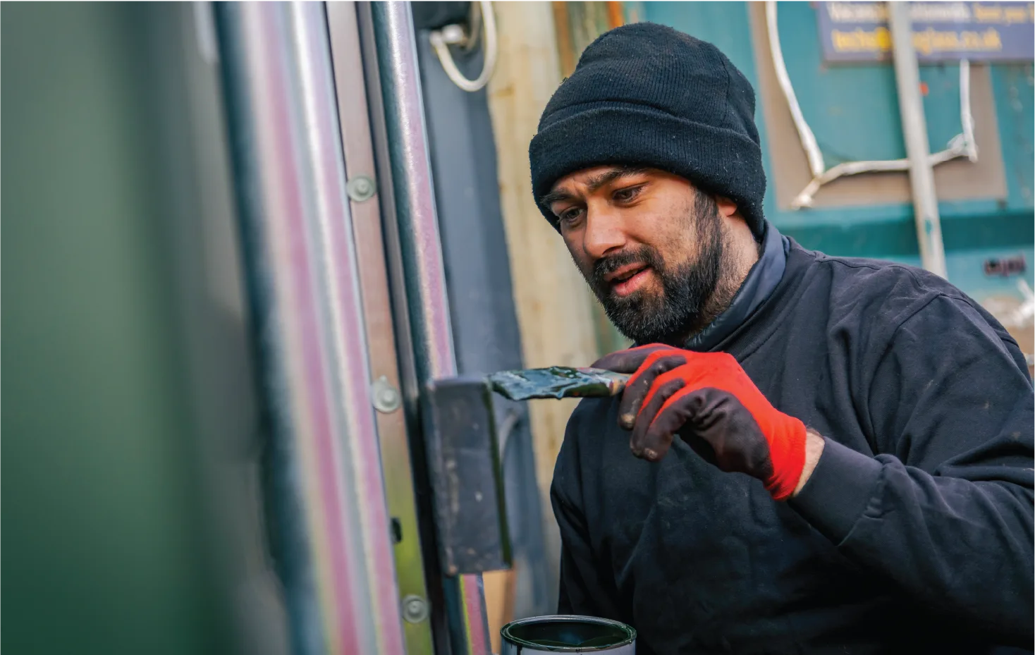 A bearded worker wearing a black beanie, black jacket, and red protective gloves is painting a metal component on a shipping container with a brush. He holds a small can of paint while focusing on applying an even coat to the surface.