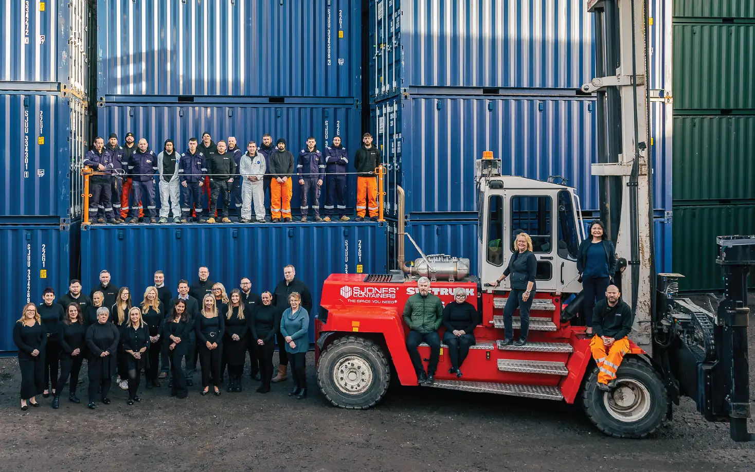 A large team photo of S Jones Containers employees posing in front of stacked blue and green shipping containers. Some team members stand on a raised platform, while others are positioned around and on a red heavy-duty forklift. The group consists of office and warehouse staff, wearing a mix of black business attire and high-visibility workwear.