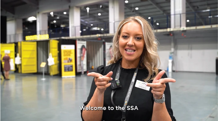 A smiling woman from S Jones Containers with blonde hair, wearing a black outfit and a lanyard with a name badge, stands in a large exhibition hall with yellow and black display booths in the background. She is gesturing with both hands while looking at the camera. The text overlay reads, 'Welcome to the SSA.'