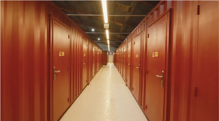 A well-lit corridor inside a storage facility made from repurposed shipping containers. The red container doors, each numbered with yellow digits, line both sides of the narrow walkway with a clean white floor.