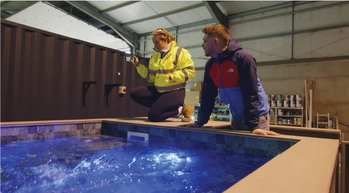 Two people inside an industrial warehouse inspecting a large container-based pool. The woman in a high-visibility yellow jacket is taking a photo, while the man in a dark blue and red jacket observes the water, which is illuminated with blue lighting.