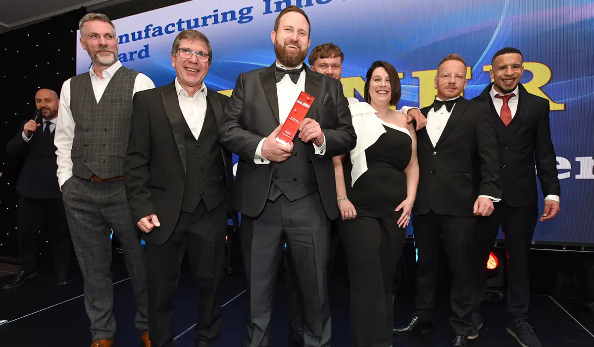 The S Jones Conversions team standing together on stage, smiling and celebrating as they hold a red trophy at the Made in the Midlands Awards 2025. The group, dressed in formal evening wear, stands in front of a large digital screen displaying the word "WINNER" in bold yellow letters. The award presenter can be seen in the background to the left. The mood is celebratory, capturing a proud moment for the team.