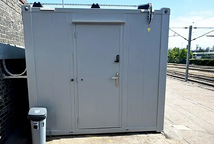 A compact, grey modular container unit positioned on a railway platform, featuring a single secure door with a lock and keypad entry. A bin is placed nearby, and railway tracks are visible in the background under a clear blue sky.