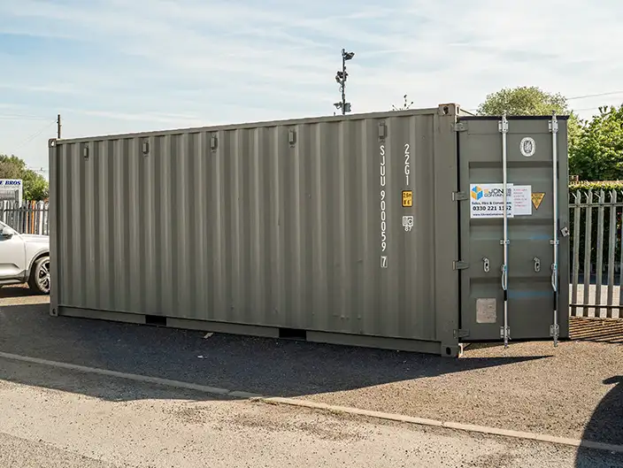 A 20ft grey shipping container positioned on a gravel surface next to a fence and parked car. The container has vertical corrugated steel sides and features a sign with the S Jones Containers logo and contact details on the front doors. The area appears to be part of an industrial or commercial lot under a clear blue sky.