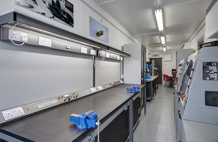 A containerised workshop fitted with workbenches, vices, electrical outlets, storage units, and specialist machinery, providing a fully equipped and organised space for technical and engineering tasks.