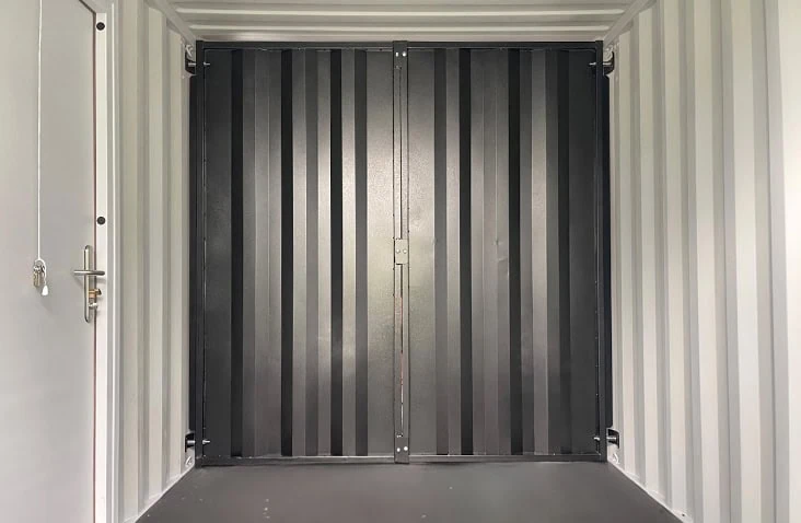 Interior view of a standard shipping container with a closed removable black steel bulkhead partition installed across the width of the unit, with light grey insulated walls and a personnel door visible on the left. 