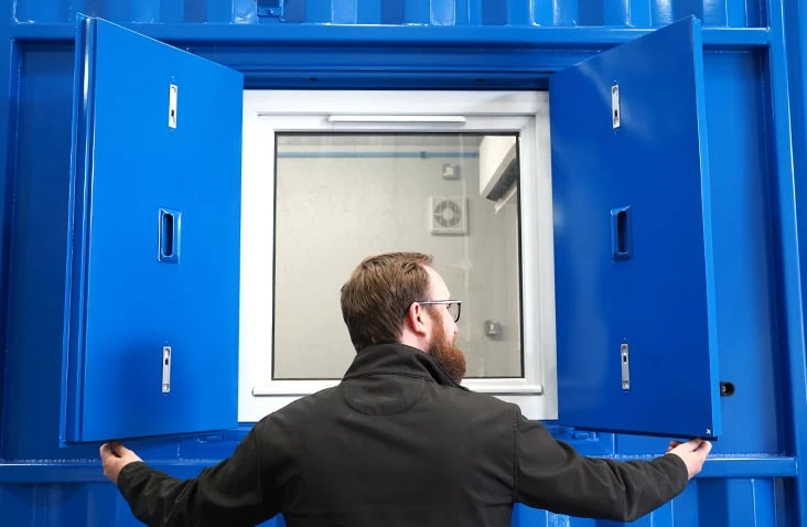 Person opening steel security shutters on a white-framed window fitted into a blue shipping container. 