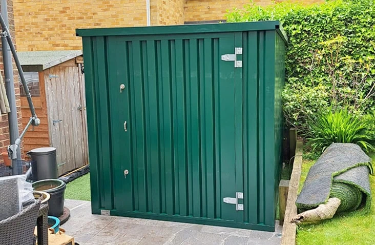 Green flat pack steel storage unit with a single door, installed in a residential garden for secure outdoor storage. 