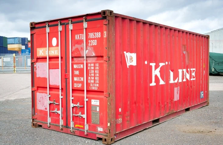     Used 20ft red shipping container with closed cargo doors, visible scuffs, rust patches, and faded branding, positioned on a yard surface with other containers in the background. 