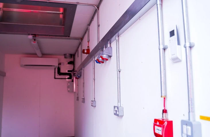 Shipping container interior fitted with surface-mounted electrical conduit, industrial sockets, fire alarm system and air conditioning unit. 