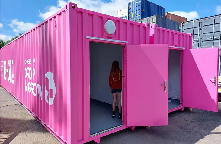 Bright pink converted shipping container with two open personnel doors, showing a person standing inside and window shutters open. 