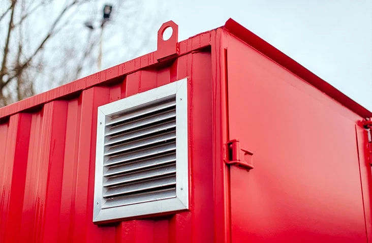 Close-up of an aluminium louvred ventilation grille mounted on the exterior of a red chemical storage container. 