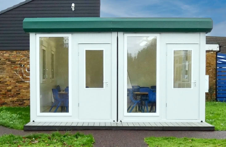 Modular two-bay classroom unit with white panelled walls, green roof trim, and large windows showing blue chairs inside, installed on a school site.