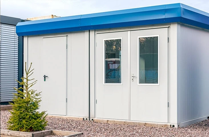 Modular two-bay office unit with white panelled walls, double-glazed doors, and a blue roof trim, set on a gravel surface with a small tree in the foreground.