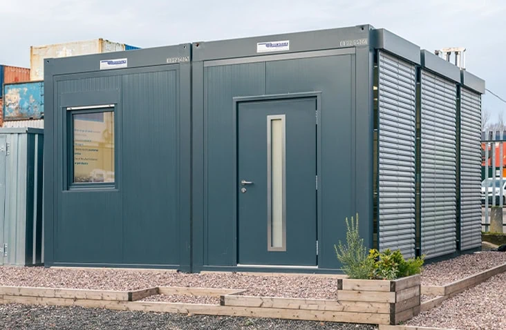Dark grey modular office unit with a central door, side window, and external metal window shutters, installed on a gravel base with wooden planters.