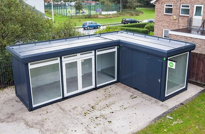 L-shaped dark grey modular building with large floor-to-ceiling windows and a single access door, installed on a concrete base beside a brick building