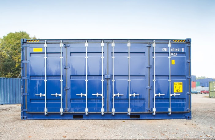     Front view of a blue 40ft high cube side opening shipping container with all side doors closed, positioned on a gravel surface in a storage yard with other containers in the background. 