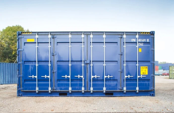    Front view of a blue 40ft high cube side opening shipping container with all side doors closed, positioned on a gravel surface in a storage yard with other containers in the background. 