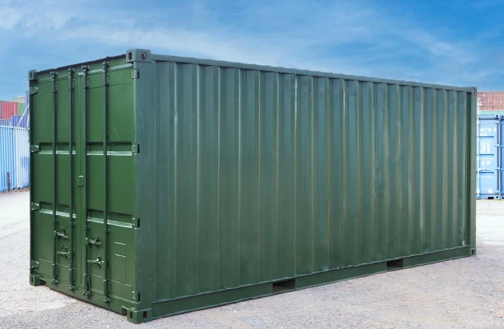 Green steel shipping container with corrugated sides and closed double doors, placed on a gravel surface outdoors with other containers visible in the background under a clear blue sky. 
