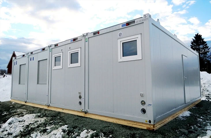 Large silver modular building made from four connected units with multiple windows and vents, installed on a raised timber base surrounded by snow