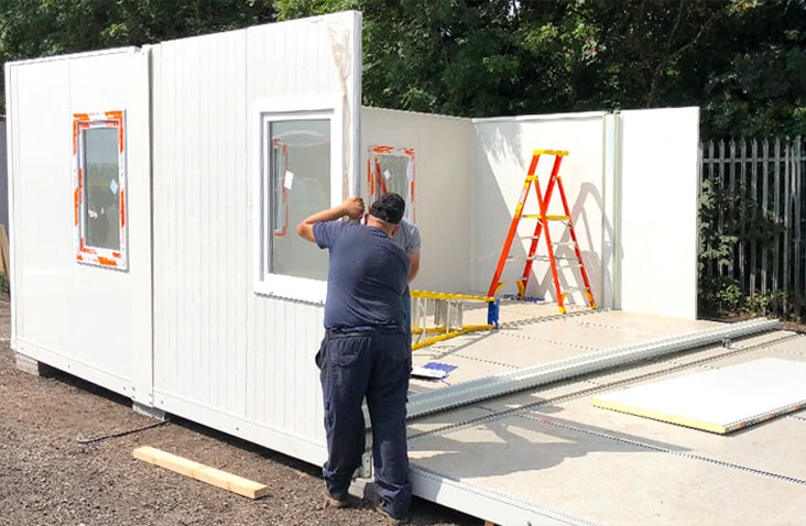 Workers assembling a flat pack office unit on site, showing panels with windows being installed during construction. 