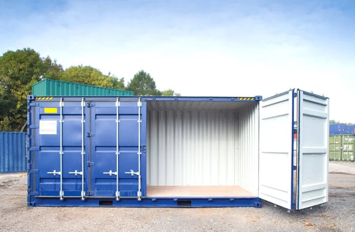 Blue 40ft high cube side opening shipping container with one side door open, revealing the light interior and wooden flooring, positioned in a storage yard with other containers and trees in the background. 