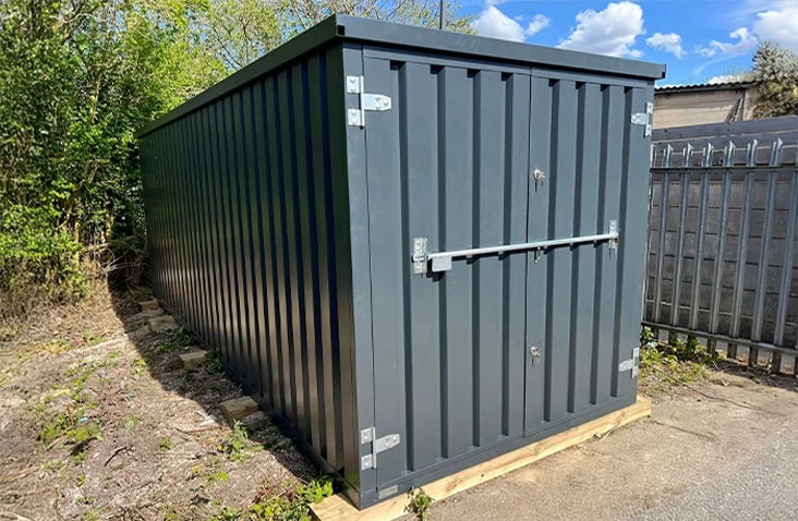     Grey flat pack steel storage unit with double doors and external locking bar, installed outdoors on timber supports. 