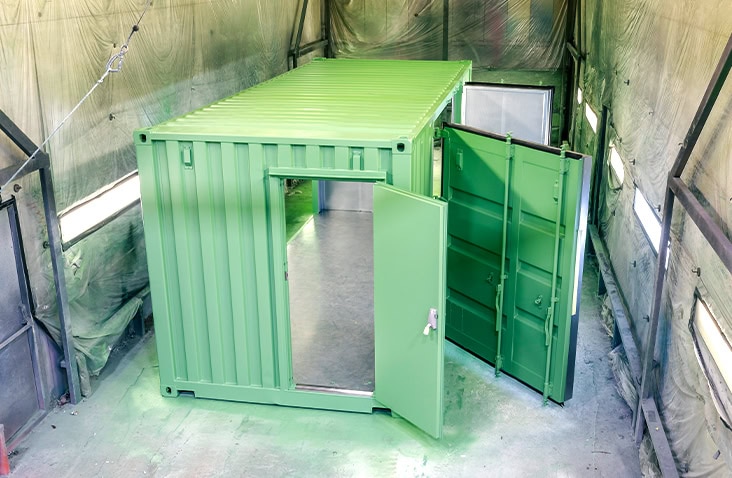 Freshly repainted light-green shipping container inside an industrial spray booth with doors open. The floor is coated in overspray paint, and protective plastic covers the booth walls. 