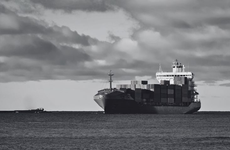 Black and white image of a cargo ship carrying shipping containers at sea, representing global shipping container transport and international freight logistics.