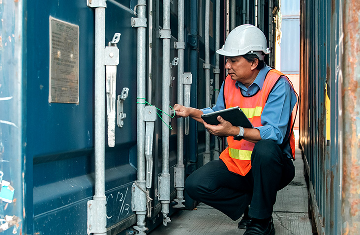 S Jones Containers specialist carrying out CSC inspection on a shipping container, checking door seals and recording details on a clipboard.