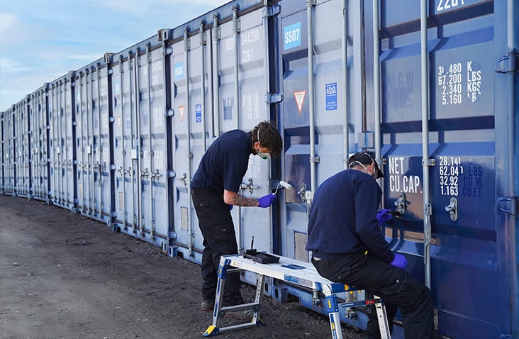 Two workers in protective masks and gloves cleaning and maintaining the locking gear on a row of blue shipping containers, using a step stool and tools on an outdoor container yard.