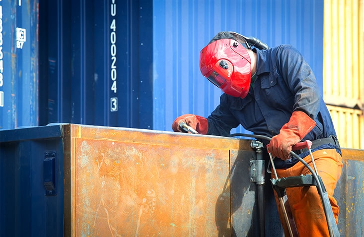 A worker wearing a red welding mask and protective gloves is using welding equipment on a steel shipping container panel, with stacked blue containers in the background. 