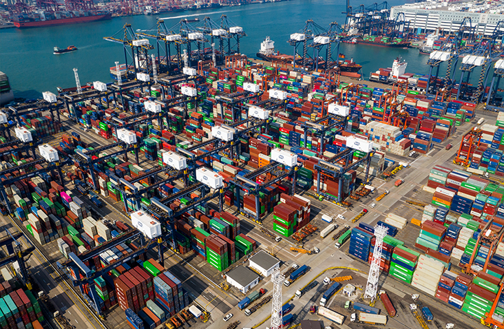 Aerial view of a large container port with thousands of shipping containers stacked in rows, cranes in operation, and vessels docked, illustrating global container logistics, port operations, and international freight handling.