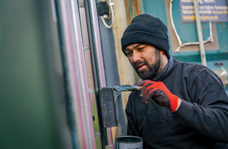 Worker wearing a hat and gloves repainting the exterior of a shipping container, applying paint to a metal surface with a brush.