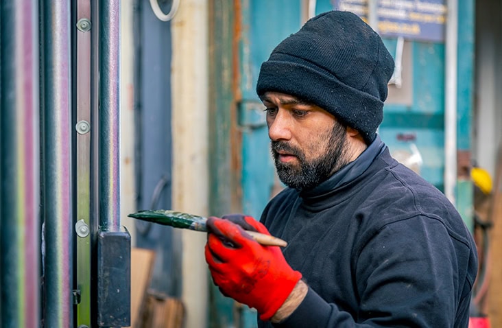 A man wearing a black beanie, black jumper, and red protective gloves carefully repainting the metal frame of a shipping container with a small brush, focusing on detail work in an outdoor workshop area.