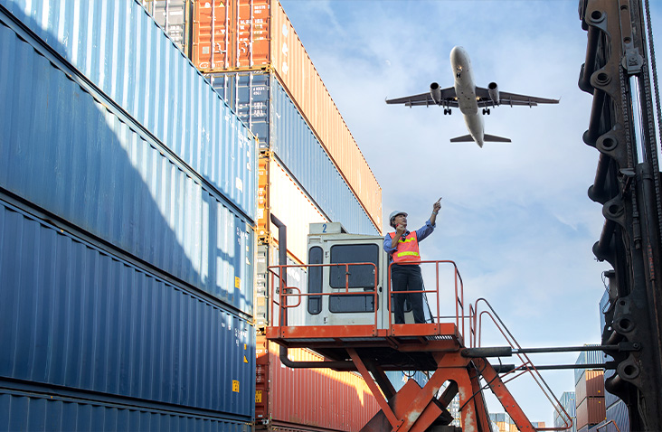 Worker on a container handler platform directing operations in a container yard, with stacked shipping containers and an aircraft flying overhead, representing integrated land and air freight logistics and global container transport.
