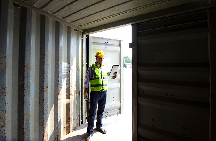 Man wearing a hard hat inspecting the interior of an open shipping container at a commercial site, holding a clipboard while checking contents, with the container doors open to daylight.
