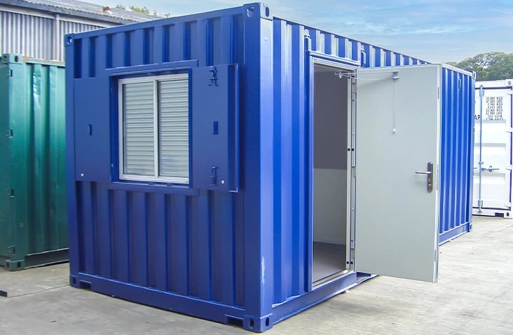 Blue 20ft converted shipping container office unit with a personnel door open and a window fitted with external steel shutters, positioned on a concrete yard.