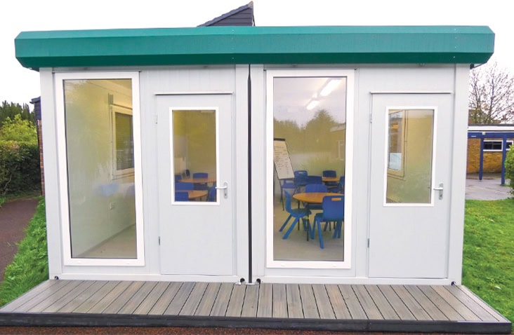 Small modular classroom building with white panelled walls, green roof trim, and large glazed doors revealing blue chairs and tables inside, installed on a wooden platform