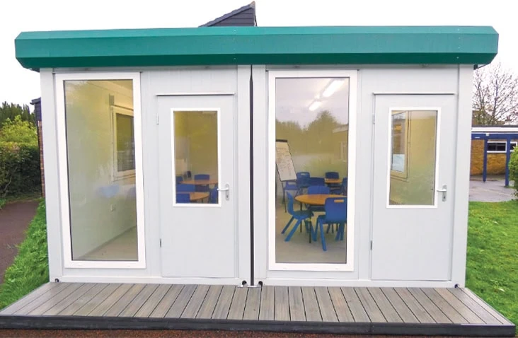 Small modular classroom building with white panelled walls, green roof trim, and large glazed doors revealing blue chairs and tables inside, installed on a wooden platform