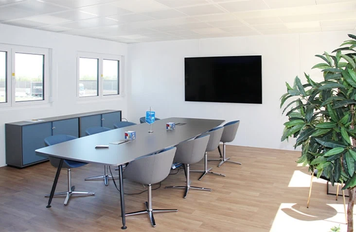 Modern modular office meeting space with a sleek table and chairs, a large wall-mounted screen, wooden flooring, and natural light from multiple windows.