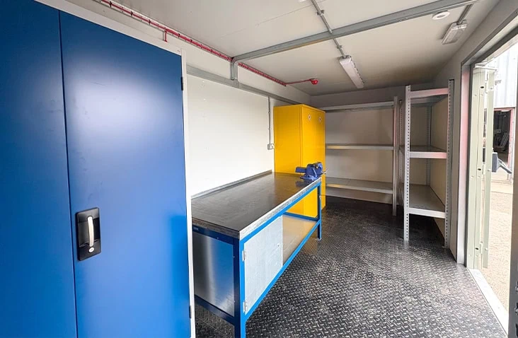 Interior of a containerised workshop featuring a metal workbench with a vice, steel floor plating, blue and yellow storage cabinets, wall-mounted power sockets, and open shelving.