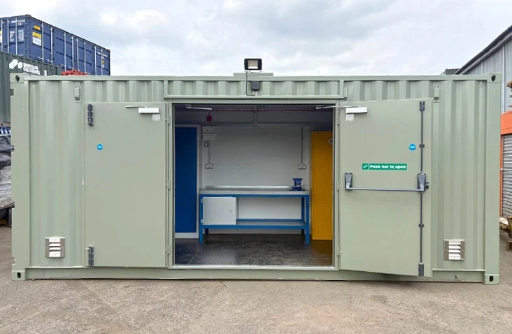 20ft converted shipping container workshop with double side-opening doors revealing an internal workbench, vice, power sockets, and storage areas. The unit is painted light green.