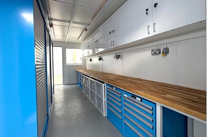 Interior of a containerised workshop fitted with a long wooden workbench, multiple tool drawers, overhead storage cabinets, power outlets along the wall, and roller-shutter storage on one side.