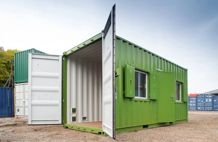 Green converted shipping container office with both cargo doors open, showing the clean white interior. The unit features two windows with external steel shutters and a personnel access door on the side, positioned on a gravel surface outdoors.