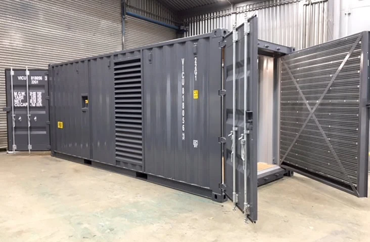 Grey containerised CSC-compliant generator housing unit with large removable louvres fitted to the cargo doors, situated inside an industrial workshop